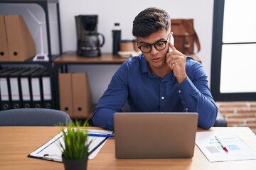 Young hispanic man business worker using laptop talking on smartphone at office