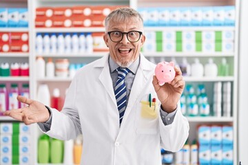 Hispanic man with grey hair working at pharmacy drugstore holding piggy bank celebrating achievement with happy smile and winner expression with raised hand