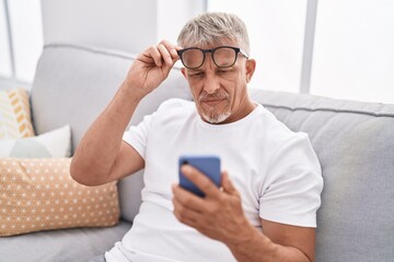 Middle age grey-haired man looking screen smartphone with vision problem at home