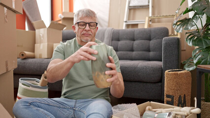 Middle age grey-haired man unpacking cardboard box sitting on floor at new home