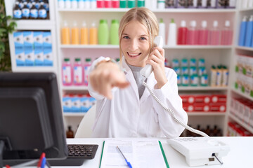 Young caucasian woman working at pharmacy drugstore speaking on the telephone pointing to you and the camera with fingers, smiling positive and cheerful