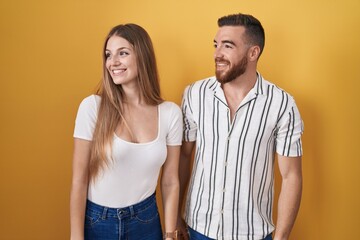 Young couple standing over yellow background looking away to side with smile on face, natural expression. laughing confident.