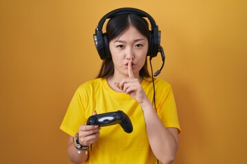 Chinese young woman playing video game holding controller asking to be quiet with finger on lips. silence and secret concept.