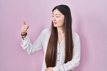 Chinese young woman standing over pink background looking proud, smiling doing thumbs up gesture to the side