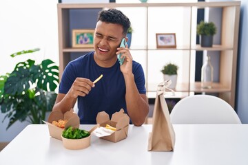 Young latin man talking on smartphone eating take away food at home