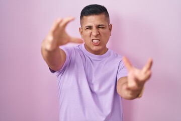 Young hispanic man standing over pink background shouting frustrated with rage, hands trying to strangle, yelling mad