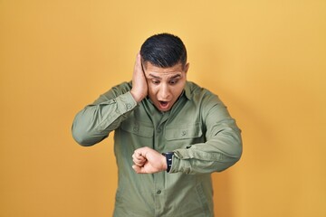 Hispanic young man standing over yellow background looking at the watch time worried, afraid of...