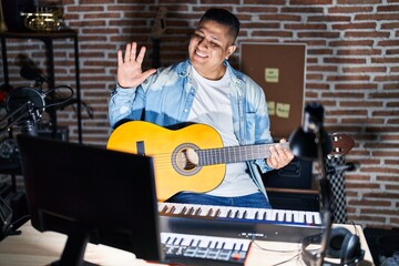 Hispanic young man playing classic guitar at music studio waiving saying hello happy and smiling, friendly welcome gesture