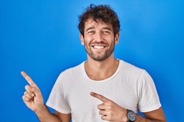 Hispanic young man standing over blue background smiling and looking at the camera pointing with two hands and fingers to the side.