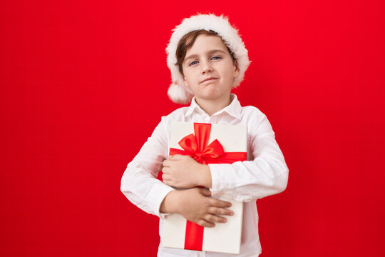 Little caucasian boy wearing christmas hat and holding gifts relaxed with serious expression on face. simple and natural looking at the camera.