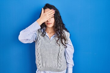 Young brunette woman standing over blue background covering eyes with hand, looking serious and sad. sightless, hiding and rejection concept