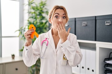 Hispanic doctor woman holding awareness orange ribbon covering mouth with hand, shocked and afraid...