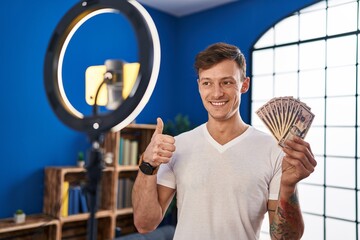 Caucasian man recording vlog tutorial with smartphone at home holding money smiling happy and positive, thumb up doing excellent and approval sign