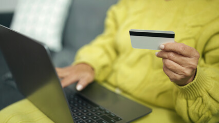 Middle age hispanic woman using laptop and credit card sitting on sofa at home