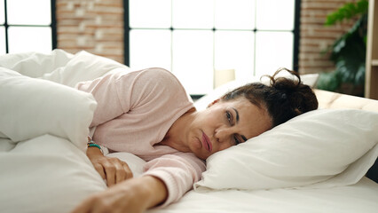 Middle age hispanic woman lying on bed with serious expression at bedroom