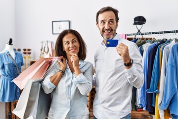 Hispanic middle age couple holding shopping bags and credit card with hand on chin thinking about question, pensive expression. smiling and thoughtful face. doubt concept.