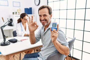 Middle age man at doctor clinic holding glucose meter device doing ok sign with fingers, smiling...