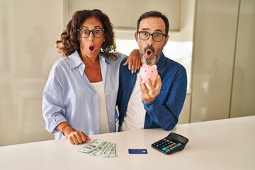 Middle age couple holding piggy bank calculating savings scared and amazed with open mouth for...