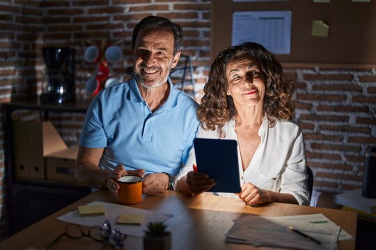 Middle Age Hispanic Couple Using Touchpad Sitting On The Table At Night Smiling Looking To The Side And Staring Away Thinking.