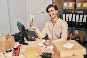 Young caucasian man ecommerce business worker holding dollars writing on notebook at office