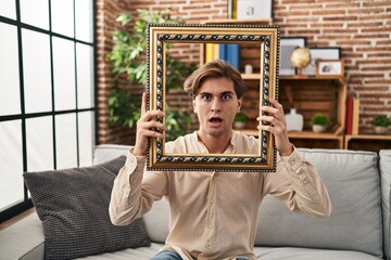 Young man holding empty frame in shock face, looking skeptical and sarcastic, surprised with open mouth