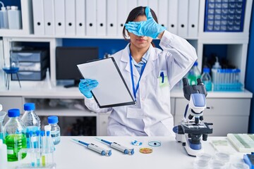 Hispanic young woman working at scientist laboratory covering eyes with hand, looking serious and sad. sightless, hiding and rejection concept