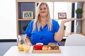 Caucasian plus size woman eating breakfast at home smiling cheerful presenting and pointing with palm of hand looking at the camera.