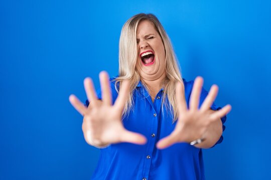 Caucasian plus size woman standing over blue background afraid and terrified with fear expression stop gesture with hands, shouting in shock. panic concept.
