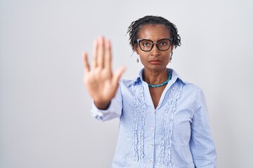 African woman with dreadlocks standing over white background wearing glasses doing stop sing with palm of the hand. warning expression with negative and serious gesture on the face.