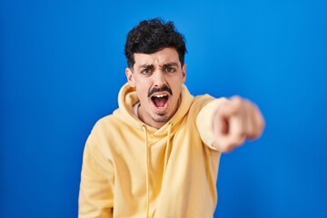 Hispanic man standing over blue background pointing displeased and frustrated to the camera, angry...