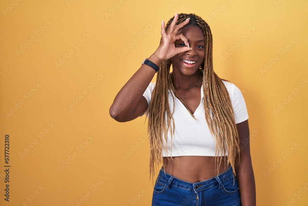 Sticker African american woman with braided hair standing over yellow background doing ok gesture with hand smiling, eye looking through fingers with happy face.