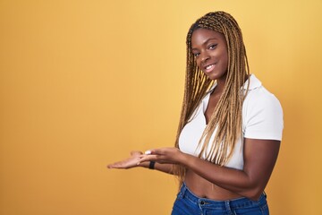 African american woman with braided hair standing over yellow background inviting to enter smiling natural with open hand