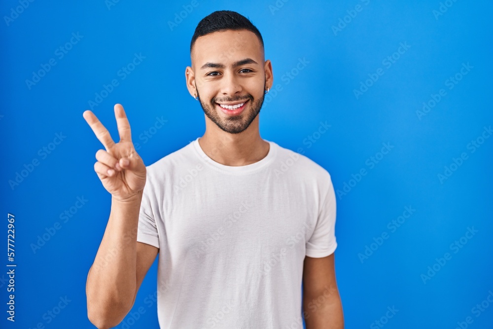 Wall mural Young hispanic man standing over blue background smiling with happy face winking at the camera doing victory sign with fingers. number two.