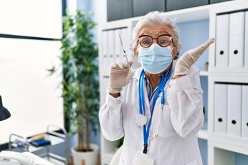 Senior woman with grey hair wearing doctor uniform and medical mask holding syringe celebrating victory with happy smile and winner expression with raised hands