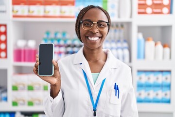 African american woman working at pharmacy drugstore showing smartphone screen looking positive and happy standing and smiling with a confident smile showing teeth