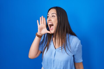 Fototapeta premium Young brunette woman standing over blue background shouting and screaming loud to side with hand on mouth. communication concept.