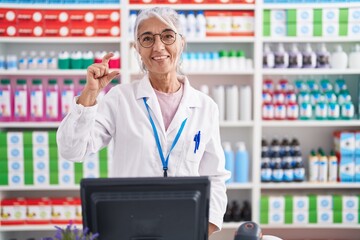 Middle age woman with tattoos working at pharmacy drugstore smiling and confident gesturing with hand doing small size sign with fingers looking and the camera. measure concept.