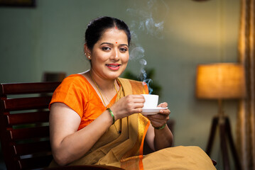 Happy smiling woman drinking tea or coffee by looking at camera while sitting on chair at home - concept of mourning routine, healthy diet and satisfaction