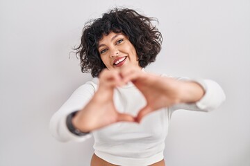 Fototapeta na wymiar Hispanic woman with curly hair standing over isolated background smiling in love doing heart symbol shape with hands. romantic concept.