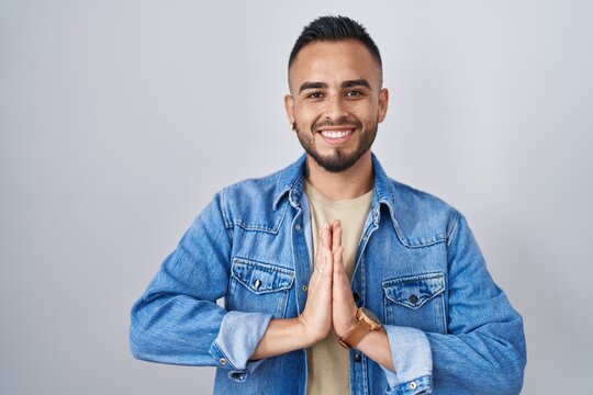 Young hispanic man standing over isolated background praying with hands together asking for forgiveness smiling confident.