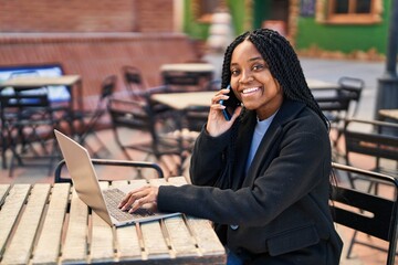 African american woman talking on smartphone using laptop sitting on table at coffee shop terrace