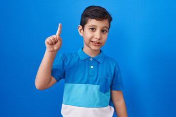 Little hispanic boy standing over blue background smiling with an idea or question pointing finger...