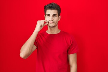 Young hispanic man standing over red background mouth and lips shut as zip with fingers. secret and...