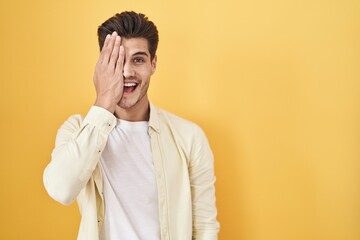 Fototapeta premium Young hispanic man standing over yellow background covering one eye with hand, confident smile on face and surprise emotion.