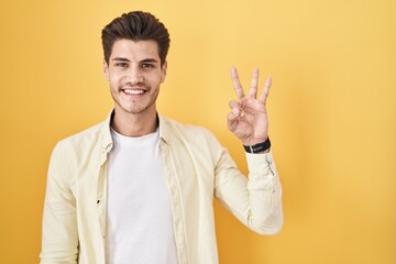 Young hispanic man standing over yellow background showing and pointing up with fingers number three while smiling confident and happy.