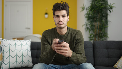 Young hispanic man watching tv sitting on sofa with boring expression at home