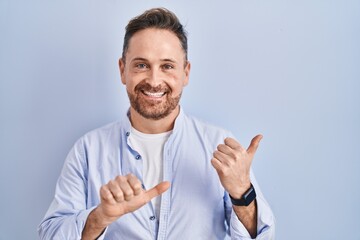 Middle age caucasian man standing over blue background pointing to the back behind with hand and thumbs up, smiling confident