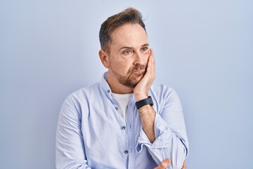 Middle age caucasian man standing over blue background thinking looking tired and bored with depression problems with crossed arms.