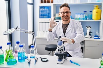 Middle age caucasian man working at scientist laboratory doing ok sign with fingers, smiling friendly gesturing excellent symbol