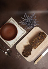 Top View Black Bread with cereals Sliced on a wooden cutting board against a brown background. Next to the bread Lies a Knife, clay plate, fork, spoon and white cotton tablecloth. Vertical shot